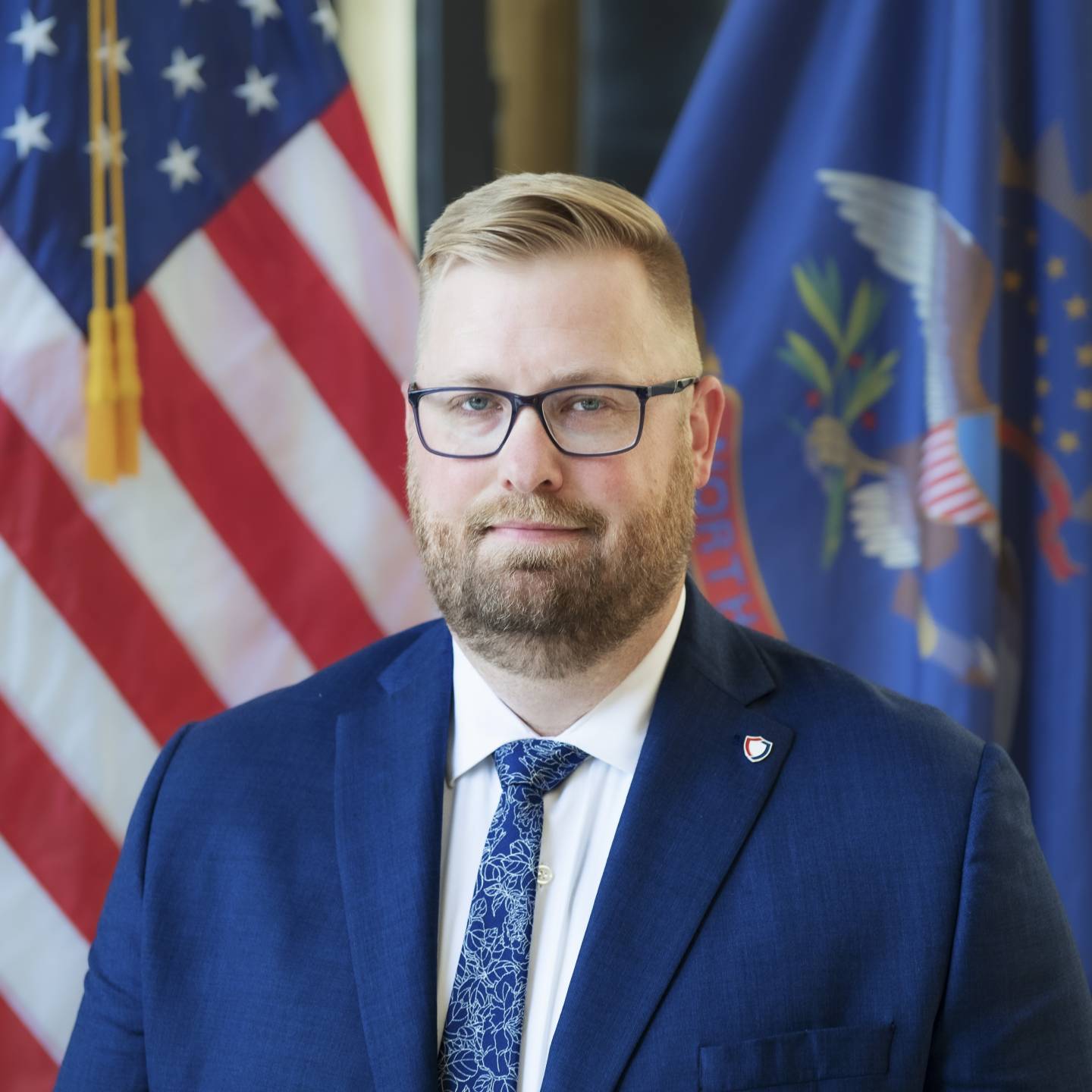 A man in a blue suit looking forward with the United States and North Dakota flags in the background.